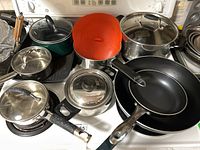 Full set of various kitchen pots, pans, and silicone lid on stove including stainless steel pots, frying pans, and a griddle pan.