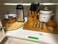 Photo showing various kitchen utensils including Henckels knife block with knives, Rival crock pot, box grater, Pyrex mixing bowl, wooden lazy Susan, wooden trivet, onion-shaped scrub brush, wooden cutting board, and slicer.