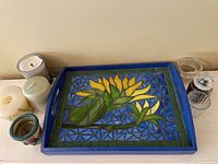 Wood tray with slag glass mosaic design of yellow flowers and green leaves bordered by blue and green geometric patterns, displayed with assorted candles and a metal decorative candle holder.