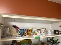 A shelf with various fine bone china mugs including floral patterns along with small china dishes and a knitted tea pot cozy.