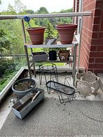 Photo showing two large terracotta planters on a white metal shelf, various small planters and plant stands below. Also visible are two decorative silver metal pots and a glass watering bulb.