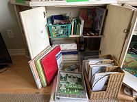 Wide view inside cabinet showing a variety of scrapbooking albums, paper sheets, a wicker paper organizer filled with paper and materials, and a plastic basket holding embellishments.