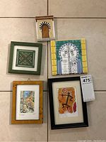 Full view of all five souvenir art pieces arranged on floor including tile, textile, cast door, and two framed paintings.