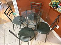 Top and angled views of a round glass dining table with green metal base and four matching green vinyl seat chairs placed around it on tiled floor.