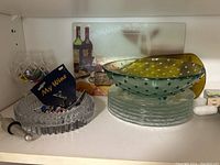 Photo showing a tempered glass cutting board with wine bottle image, a green blown glass bowl with dimples, clear blown glass plates, a crystal cut glass bowl, and wine glass charms
