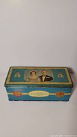 Rectangular blue lithographed tin with images of Queen Elizabeth II and Prince Philip on the lid, photographed from the front.