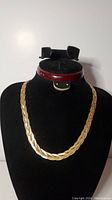 Full view of the gold coloured braided necklace displayed on a black velvet neck form with a measuring tape case in the background.