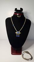 Front view of necklace on black neck form, showing silver beads, lapis lazuli beads, and blue inlaid capsule pendant with hanging teardrop elements. Snake bracelet placed adjacent on surface.