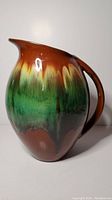 Full view of ceramic jug showing brown, cream, and green drip glaze with handle and spout.