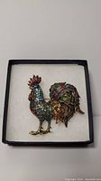 Front view of multicolored crystal-encrusted rooster brooch in box, showing full figure with vibrant stones.