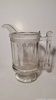 Full view of the clear glass pitcher showing the spout, handle, and body with vertical panels and textured star pattern bands.