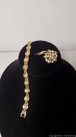 Full view of gold colored rhinestone bracelet alongside matching brooch displayed on black neck form.