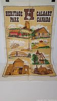 Full front view of the linen tea towel showing multiple illustrated scenes from Heritage Park and the text 'Heritage Park Calgary Canada' printed at the top.
