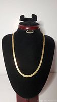 Full view of gold coloured herringbone flat chain necklace displayed on a black neck form.