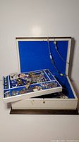 Closed cream jewellery box with blue interior, containing mixed jewellery items including bracelets, necklaces, earrings.