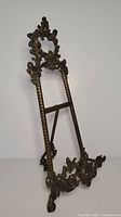 Front angle view of the ornate bronze-colored easel showing intricate scrollwork and floral motifs.