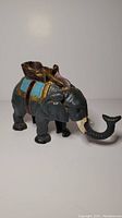 Side profile of painted cast iron elephant coin bank with visible blue saddle cloth and rider basket on top.