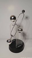 Front view of the perpetual motion desk sculpture showing one large and two smaller polished metal spheres suspended in motion on metal wires supported by clear acrylic stands on a black base.