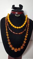 Three necklaces displayed on a black neck form: large round amber beads, yellow-orange beads, and brown elongated beads strand.
