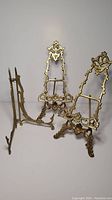 Photo showing all three brass stands: two ornate easels and one folding plate stand with intricate scrollwork and details.