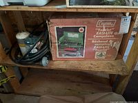 Original boxed Coleman camp stove model 431, green with red component, visible on shelf with hoses and propane tanks