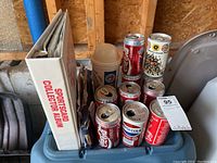 White sports card collector album with baseball cards and loose cards inside. Assorted collectible beverage cans including Coca-Cola and Pepsi. Plastic container with stamps. Items stored in a garage environment.