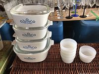 Four stacked Corning Ware lidded casserole dishes with blue floral design and seven white ramekin bowls to the side.