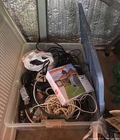 Clear tote with electrical cords, cables, power strips, and boxed outdoor socket device inside.