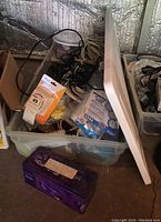 Clear Rubbermaid plastic tote with lid partially opened showing a mix of cables and adapters inside, purple metal tin in front