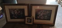 Three framed floral prints together on floor: two large matching frames and one small frame in front; small frame has bio on back, one large frame shows damage