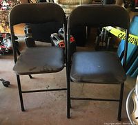 Two black folding chairs placed side by side showing both front views and seats with visible marks.