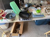 Wide view of a table with various kitchen ware items including bakeware pans, plastic containers, utensils, bowls, and more.