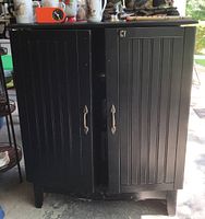 Front view of the black media cabinet showing double swing out doors with vertical grooves and decorative handles, placed on short legs.