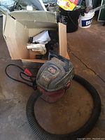 3.8 litre red and black Micro brand shop vacuum with hose and attachments in box beside it on garage floor