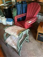 Red resin Muskoka chair with arm fading and metal foot rests with fabric cushions showing wear and scratches in garage setting.