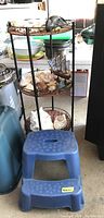 Full view of the metal framed shelf with three wicker wrapped woven levels, round shape, with various items on shelves (not included). Blue two-step plastic stool with textured steps in front of shelf.