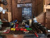 Wide view of table with assorted hand tools including screwdrivers, hammers, funnels, hardware organizer with labeled drawers, and metal tools arranged on surface inside garage.