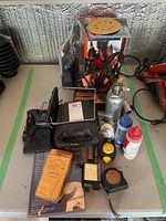 Photo showing Motomaster Eliminator charger/inverter, B&D sander, hydraulic jack, stud finder, chalk bottles, tape measures, and small tools on table.