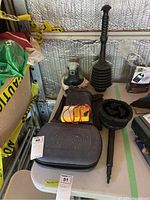 Photo of Mastercraft electric scissors case, black rubber plunger with wooden handle, black brush hose attachment, and handheld battery tester on shelf with other items