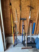 Full view of yard tools lined up against plywood wall in garage including rakes, snow shovels, broom, spade, and telescoping pole.