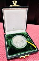 Coin in original black presentation box with green velvet interior and white satin lid lining, showing the coin secured inside a clear capsule.