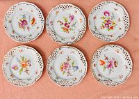 Six antique Dresden porcelain plates arranged on a pink background showing floral patterns and pierced lattice edges with gold trim.