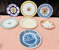 Seven antique decorative plates arranged on a pink fabric surface, showing various floral, nautical, and heraldry designs from a forward angle.