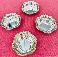 Four polished sterling silver nut dishes with scalloped and floral decorated edges placed on a red textured cloth