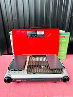 Front view of Coleman camp stove with red lid open, showing the grill and side wind guards. Original box visible behind.