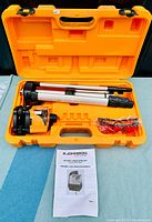 Open yellow plastic case showing rotary laser level, folded tripod, laser attachments in plastic, and manual.