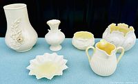 Lot showing six porcelain pottery pieces: five Belleek items including a cream-colored vase with raised floral design, star candy dish, cabbage leaf bowl, lotus sugar bowl, double spout creamer; and a white Lenox bud vase with gold trim.