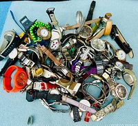 Photo showing a large pile of assorted wristwatches with different band types and colored faces on a light blue surface.