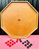 Full view of the vintage wooden crokinole game board, showing octagonal shape, painted scoring circles and original red and black game pieces arranged below it.
