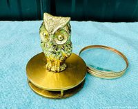 Solid brass owl figurine on circular brass base with magnifying glass loupe swung out to the side. The item is photographed on a cloth surface with a dark backdrop.